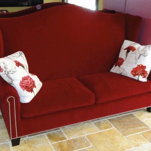 May include: A red velvet loveseat with a high back and two floral throw pillows. The pillows feature red flowers on a white background. The loveseat has a studded trim along the arms and base.