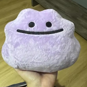 May include: A plush, lavender-colored Ditto Pokémon character with a smiling face. The stuffed toy has two black oval eyes and a curved black mouth. The plush is held in a person's hand.