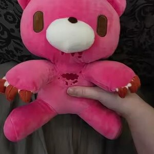 May include: A pink plush bear with brown accents around the eyes and ears, a white muzzle, and brown claws. The bear has a distressed design with red accents. The bear is being held by a person.