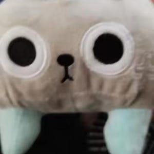 May include: A soft, plush toy resembling a creature with large black eyes, a small black nose, and light blue feet. The toy is light grey in colour and appears soft and cuddly.