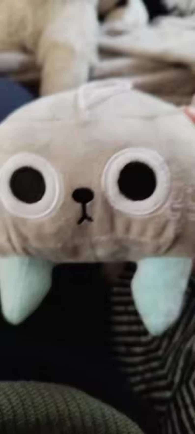 May include: A soft, plush toy resembling a creature with large black eyes, a small black nose, and light blue feet. The toy is light grey in colour and appears soft and cuddly.