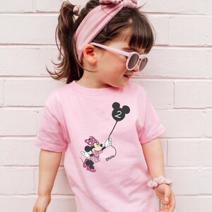 A young girl wearing a pink t-shirt with a Minnie Mouse design and sunglasses is leaning against a white brick wall.