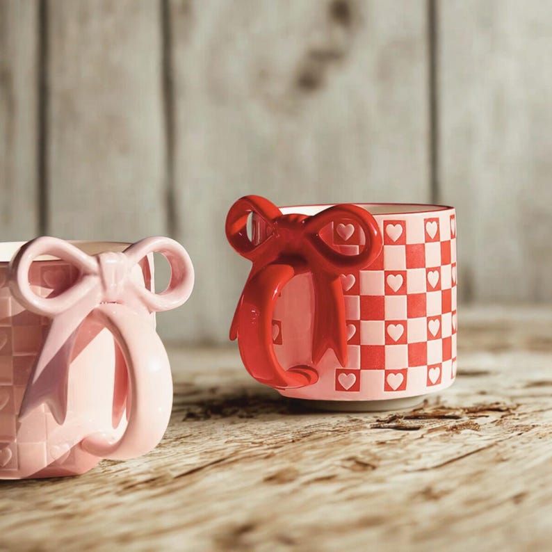 May include: Two ceramic mugs with bow-shaped handles. One mug is light pink with a bow handle, and the other is red with a bow handle. The red mug has a checkered pattern with white hearts. The mugs are on a wooden surface.