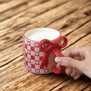 May include: A ceramic mug with a red bow-shaped handle. The mug features a red and white checkered pattern with heart accents. The mug is filled with a white liquid.