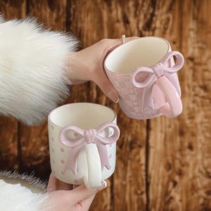 May include: Two ceramic mugs with pink bow handles. One mug has a pink polka dot pattern, and the other has a pink basketweave design. Both mugs have a white interior and are held against a wooden background.