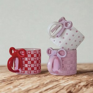 May include: Three decorative ceramic mugs stacked, featuring bow-shaped handles. One mug has a red and white checkered pattern with heart accents, while another has a pink checkered pattern with heart accents. The third mug is white with pink polka dots.