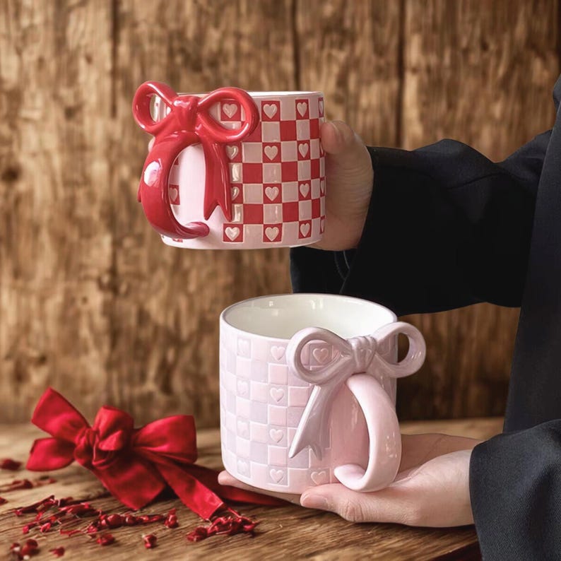 May include: Two ceramic mugs with bow-shaped handles. One mug is red and white checkered with heart accents, the other is pink with a similar pattern. The mugs are held up against a wooden background.