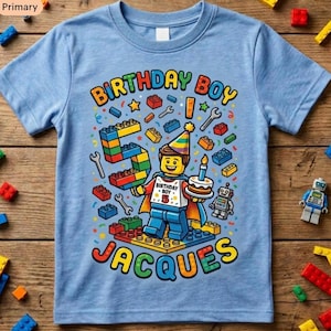 May include: Light blue short-sleeved t-shirt with a colorful graphic. The graphic features the text "BIRTHDAY BOY" and "JACQUES", along with a Lego-style figure holding a cake and the number "5". The design includes Lego bricks, wrenches, and a robot.