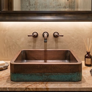 Puede incluir: Un lavabo cuadrado de cobre martillado con una base de pátina verde azulado sobre una encimera de mármol beige. El lavabo cuenta con un grifo de bronce montado en la pared con tres manijas. Incluye una pastilla de jabón, difusor y toallas.
