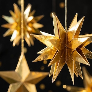 May include: Multiple three-dimensional gold star ornaments crafted from folded paper with a metallic finish. Each star features numerous sharp, pointed rays and is suspended by a thin gold string against a dark background.