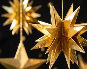 May include: Multiple three-dimensional gold star ornaments crafted from folded paper with a metallic finish. Each star features numerous sharp, pointed rays and is suspended by a thin gold string against a dark background.