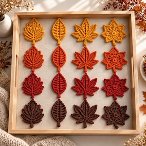 Può includere: Una collezione di sedici foglie all'uncinetto disposte in una griglia quattro per quattro all'interno di un vassoio in legno chiaro. Le foglie presentano varie forme e colori ispirati all'autunno, tra cui giallo, arancione, rosso e marrone.