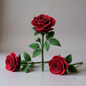 May include: Three handmade crochet roses in vibrant red, with green stems and leaves. One rose stands upright, while the other two lie horizontally. The detailed crochet work creates a realistic floral appearance, perfect for home decor or gifting.