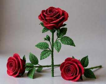 Könnte beinhalten: Drei handgefertigte Häkelrosen in leuchtendem Rot, mit grünen Stielen und Blättern. Eine Rose steht aufrecht, die anderen beiden liegen horizontal. Die detaillierte Häkelarbeit erzeugt ein realistisches Blumenaussehen, ideal für Heimdekoration oder als Geschenk.