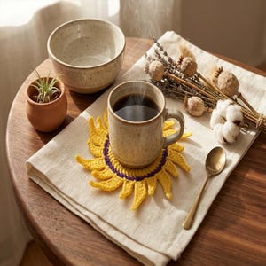 May include: A ceramic mug filled with dark coffee sits on a yellow and purple crocheted sunflower coaster. The coaster rests on a white linen cloth alongside a small metal spoon, a terracotta pot with a small air plant, a ceramic bowl, and dried flowers.
