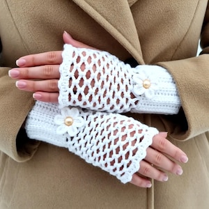 Könnte beinhalten: Ein Paar weiße Häkel-Fingerhandschuhe. Die Handschuhe haben ein rautenförmiges Lochmuster und sind mit einer weißen Blume und einer goldfarbenen Perle verziert. Die Handschuhe werden vor einem beigefarbenen Mantel gezeigt.