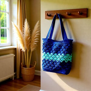 May include: A blue tote bag with a textured design and a turquoise stripe. The bag is hanging on a wooden wall hook. The bag has two handles and is made of a woven material. The bag is a medium size.