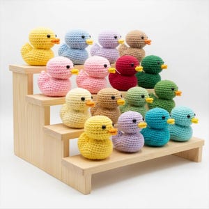 Könnte beinhalten: Eine Sammlung bunter, gehäkelter Entenküken, die auf einem gestuften Holzregal ausgestellt sind. Die Enten sind in verschiedenen Farbtönen gehalten, darunter Gelb, Rosa, Blau, Grün und Braun, jede mit gelbem Schnabel und schwarzen Augen. Ein charmantes Deko-Objekt.