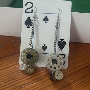 May include: Unique handmade earrings featuring silver-tone chain and ear hooks. Each earring showcases a repurposed sewing machine bobbin, adorned with small buttons. The earrings are displayed against a playing card with the number 2 and spades.