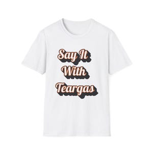 Puede incluir: Camiseta blanca con el texto "Say It With Teargas" en una fuente retro. El texto es marrón con un contorno negro, creando un efecto 3D. La camiseta es de estilo cuello redondo clásico.