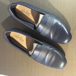 May include: A pair of navy blue leather loafers. The shoes have a textured band across the top and the brand name "collection by Clarks" is visible inside. The shoes are new and unworn.