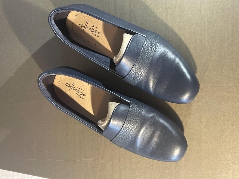 May include: A pair of navy blue leather loafers. The shoes have a textured band across the top and the brand name "collection by Clarks" is visible inside. The shoes are new and unworn.