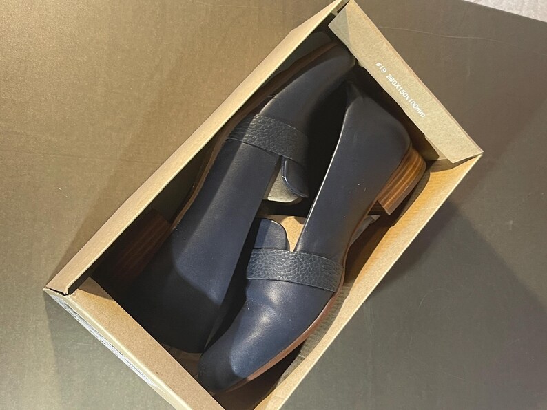 May include: A pair of navy blue leather loafers with a textured strap detail, presented inside a cardboard box. The shoes have a low wooden heel and a classic design, suitable for various occasions. The box has text that reads "#19 280x160x100mm".