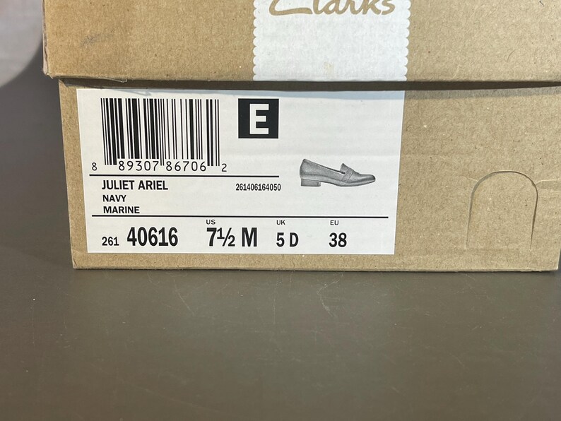 May include: A brown cardboard shoe box with a label displaying product information. The label includes the text "JULIET ARIEL NAVY MARINE", a barcode, and shoe size details: US 7 1/2 M, UK 5 D, and EU 38. A small illustration of a shoe is also present.