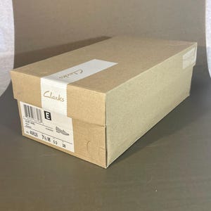 May include: A rectangular Clarks shoe box in shades of brown. The box has a white label with the brand name and shoe details. The size is 7 1/2 M, and the European size is 38.
