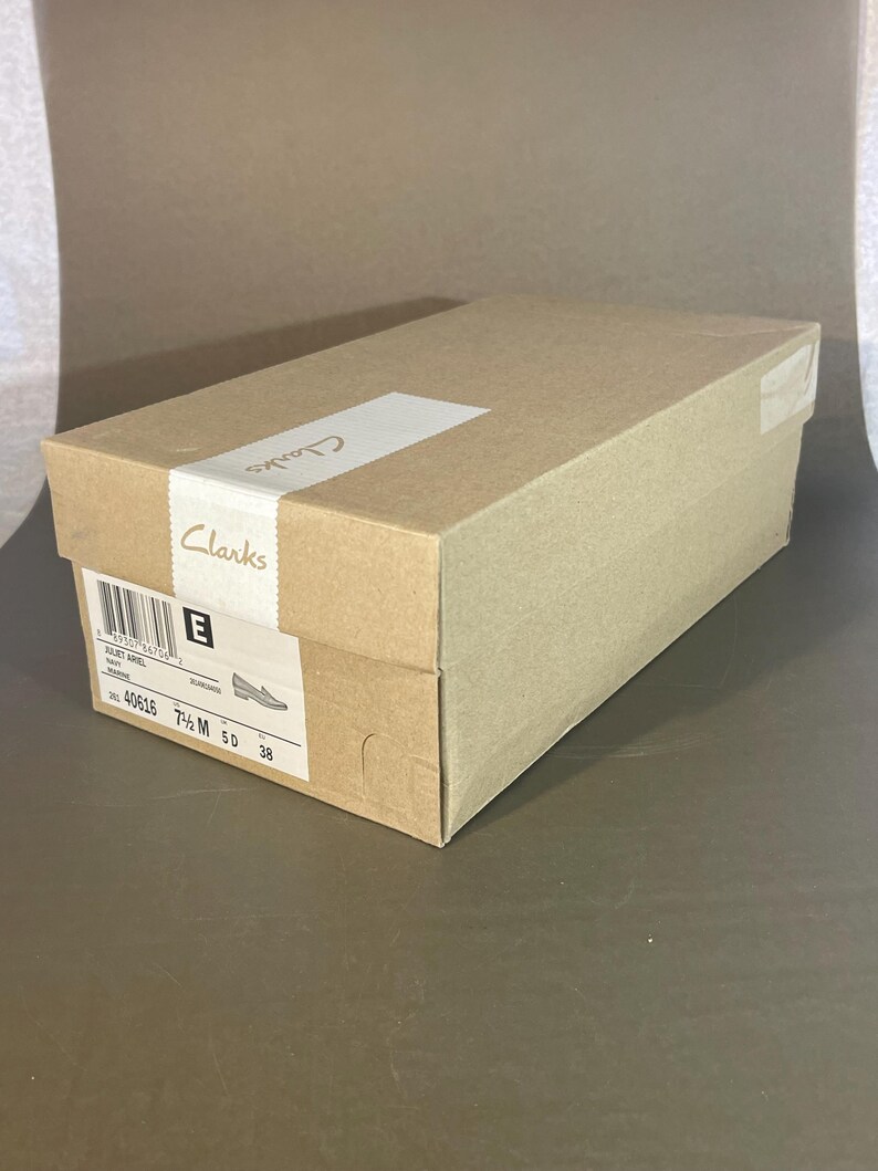 May include: A rectangular Clarks shoe box in shades of brown. The box has a white label with the brand name and shoe details. The size is 7 1/2 M, and the European size is 38.