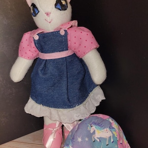 Puede incluir: Muñeca de gato blanco con una blusa rosa con lunares y un vestido de mezclilla con ribete de encaje blanco. La muñeca tiene zapatillas de ballet rosas y una bolsa a juego con temática de unicornio. El gato tiene ojos azules y orejas naranjas.