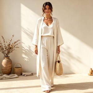May include: A cream-colored three-piece outfit consisting of a sleeveless V-neck top, wide-leg trousers with an elastic waistband, and a long, open-front duster cardigan with wide sleeves. The set is made of a textured, lightweight fabric.