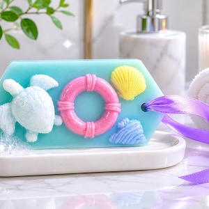 May include: A hexagonal light blue soap bar featuring a white sea turtle, a pink life preserver, a yellow seashell, and a light blue seashell. The soap has a hole on the right side with a purple satin ribbon threaded through it.