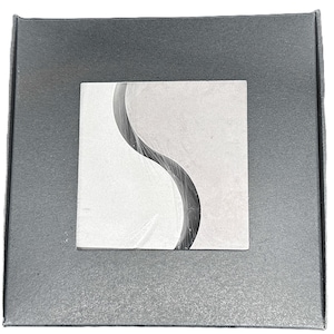 Puede incluir: Un objeto cuadrado gris claro con un diseño curvo gris oscuro, colocado sobre una caja cuadrada gris más oscura. El objeto parece estar envuelto en plástico transparente. La estética general es minimalista y moderna.