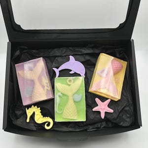 May include: A black gift box with a clear lid, filled with mermaid-themed soaps and decorative accents. The soaps feature mermaid tail designs in pink, yellow, and green. Additional sea-themed shapes include a starfish, seahorse, and dolphin.