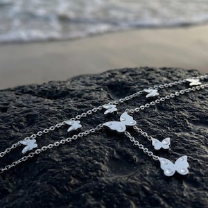 May include: A silver butterfly necklace with multiple butterfly pendants. The necklace is displayed on a dark, textured surface with a blurred ocean background. The butterflies are detailed and appear to be made of metal.