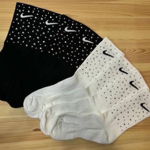 May include: Two pairs of athletic socks, one black and one white, with the Nike swoosh logo. The black socks have small, sparkling embellishments. The white socks also have the Nike swoosh logo and small embellishments.