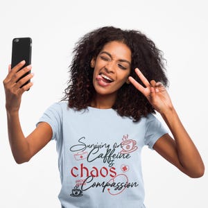May include: A light blue t-shirt with the words "Surviving on Caffeine, Chaos, Compassion" in black and red script. The shirt features coffee cup and heart graphics. A person is holding a black smartphone and making a peace sign.
