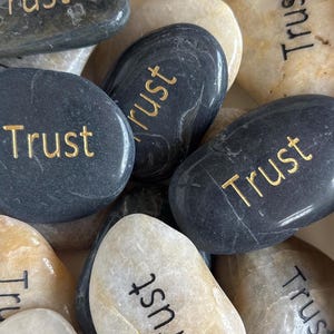 May include: A collection of smooth, polished river stones in shades of black, gray, and beige. Each individual stone features the word Trust printed in either black or gold lettering. The stones are arranged in a dense, overlapping cluster.