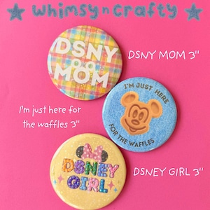 Pode incluir: Três botões redondos coloridos de 7,6 cm com texto e gráficos em um fundo rosa. Um botão diz "DSNY MOM" em um fundo xadrez. Outro diz "I'm just here for the waffles" com o Mickey Mouse de waffle. O terceiro diz "DSNEY GIRL".