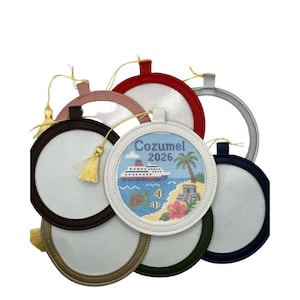 May include: A collection of circular ornaments with leather-like frames in various colors, including white, red, black, silver, and tan. One white ornament features a cross-stitch design of a cruise ship, palm tree, and sea life with the text Cozumel 2026.