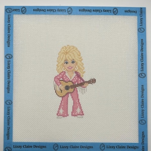 May include: A needlepoint canvas featuring a blonde figure with voluminous hair wearing a pink jumpsuit with white star accents, playing a brown acoustic guitar. The canvas is bordered by blue tape printed with the text Lizzy Claire Designs.
