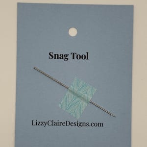 May include: A snag tool on a light blue card with the text "Snag Tool" and "LizzyClaireDesigns.com". The tool is silver with a needle-like point and a textured handle. A piece of patterned tape secures the tool to the card.
