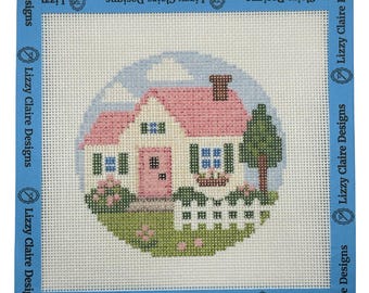 May include: A square needlepoint canvas featuring a circular design of a house with a pink roof, a pink door, and green shutters. A white picket fence, a green tree, and pink flowers are in the foreground. The blue border is printed with Lizzy Claire Designs.