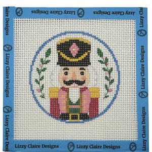 May include: A needlepoint canvas featuring a colorful nutcracker design. The nutcracker wears a black hat with a pink jewel, a pink jacket, and a green vest. The design is framed by a blue border with the text "Lizzy Claire Designs".