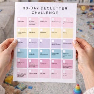 Op de afbeelding: Een afdrukbare 30-daagse declutter challenge checklist met kleurrijke vakken voor verschillende taken. De tekst "Download on Etsy" staat op een rode banner. De afbeelding wordt door een persoon vastgehouden.