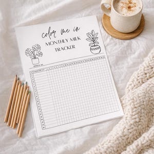 Op de afbeelding: Een witte maandelijkse melktracker met de tekst "Color Me In" en plantillustraties. Een kopje koffie staat op een houten onderzetter. Een bundel kleurpotloden ligt aan de linkerkant van de tracker.