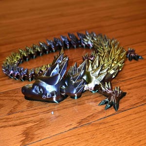 May include: A 3D-printed articulated dragon toy in iridescent colors of blue, purple, and gold. The dragon is posed on a wooden surface, showcasing its detailed scales and spiky spine. A unique decorative item.
