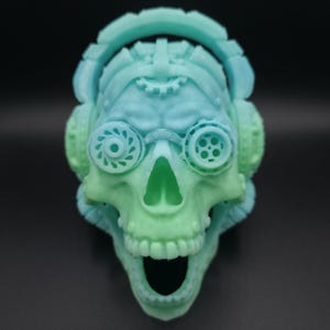 May include: A two-toned skull sculpture in shades of blue and green, featuring steampunk-inspired goggles and headphones. The skull has an open mouth and detailed teeth. The design incorporates gear-like elements.