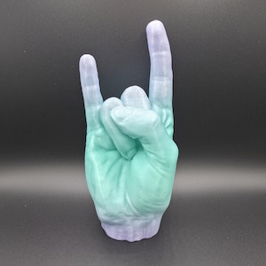 May include: A 3D-printed hand sculpture in the 'rock on' gesture. The sculpture is a gradient of colors, transitioning from a light teal to a lavender hue. The hand is detailed and textured, with a matte finish, and sits on a dark gray surface.